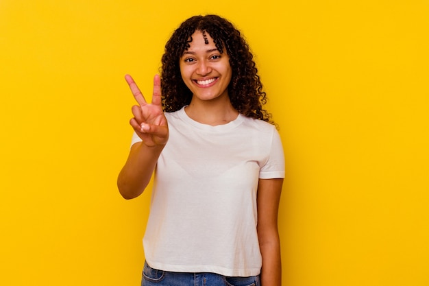 Jeune femme métisse isolée sur fond jaune montrant le signe de la victoire et souriant largement.