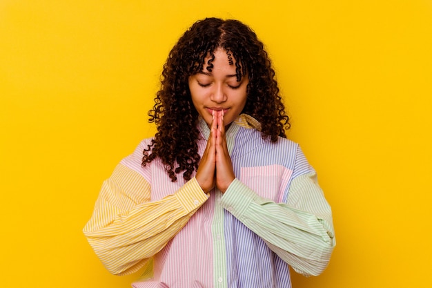 Jeune femme métisse isolée sur fond jaune priant, montrant la dévotion, personne religieuse à la recherche d'inspiration divine.