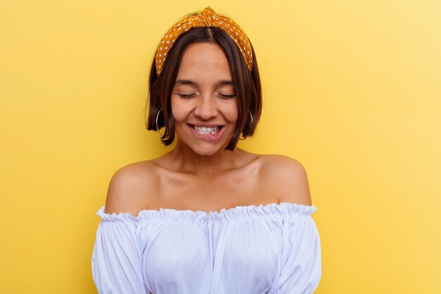 Jeune femme métisse isolée sur fond jaune rit et ferme les yeux, se sent détendue et heureuse.