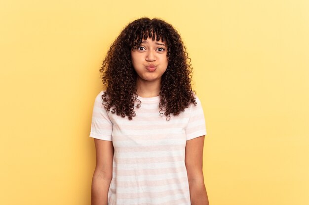 Jeune femme métisse isolée sur fond jaune souffle les joues, a une expression fatiguée. Concept d'expression faciale.