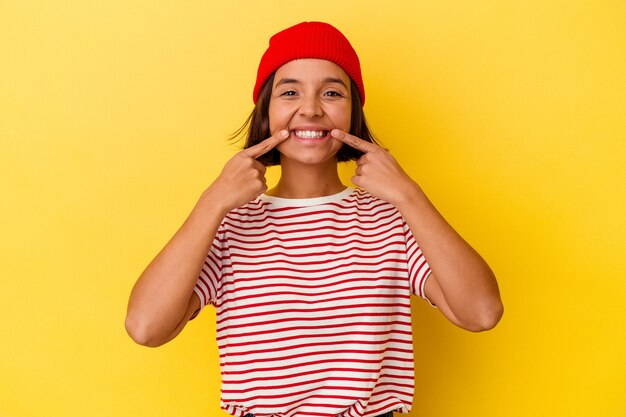 Jeune femme métisse isolée sur fond jaune sourit, pointant du doigt la bouche.