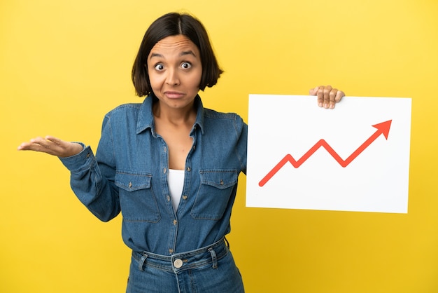 Jeune femme métisse isolée sur fond jaune tenant une pancarte avec un symbole de flèche de statistiques croissante ayant des doutes