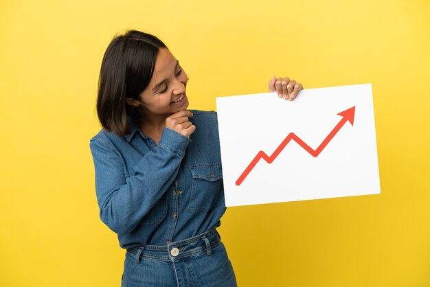 Jeune femme métisse isolée sur fond jaune tenant une pancarte avec un symbole de flèche de statistiques croissantes et pensant