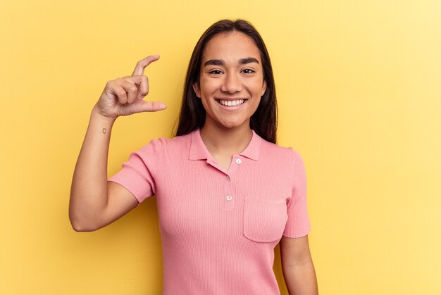 Jeune femme métisse isolée sur fond jaune tenant quelque chose de petit avec les index, souriante et confiante.