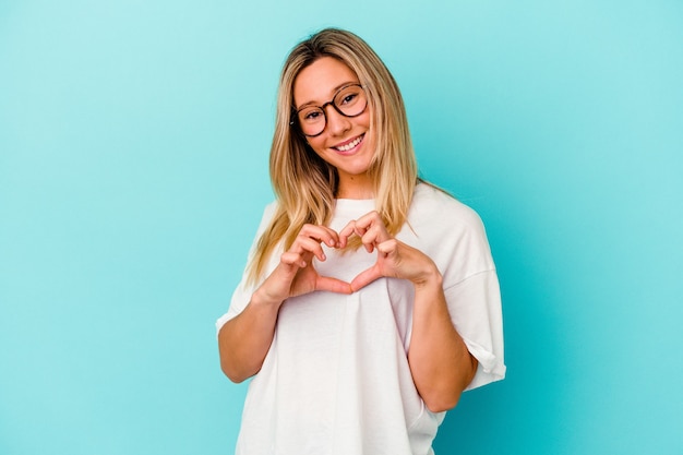 Jeune femme métisse isolée sur mur bleu souriant et montrant une forme de coeur avec les mains.