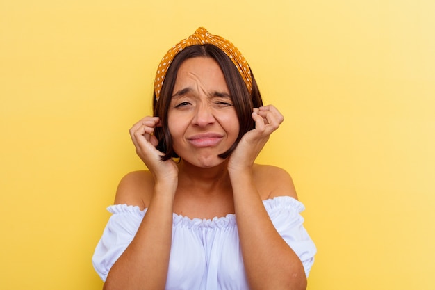 Jeune femme métisse isolée sur un mur jaune couvrant les oreilles avec les mains.