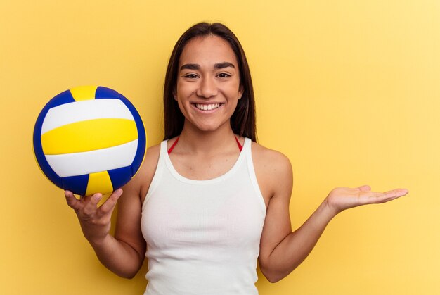 Jeune femme métisse jouant au volley-ball sur la plage isolée sur fond jaune montrant un espace de copie sur une paume et tenant une autre main sur la taille.