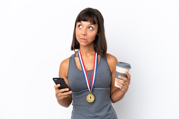 Jeune femme métisse avec des médailles isolées sur fond blanc tenant du café à emporter et un mobile tout en pensant à quelque chose