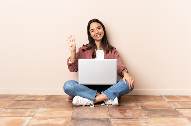 Jeune femme métisse avec un ordinateur portable assis sur le sol heureux et en comptant quatre avec les doigts