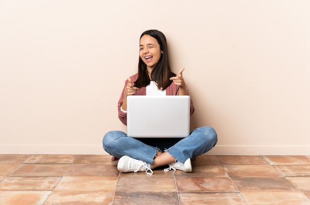 Jeune femme métisse avec un ordinateur portable assis sur le sol pointant vers l'avant et souriant