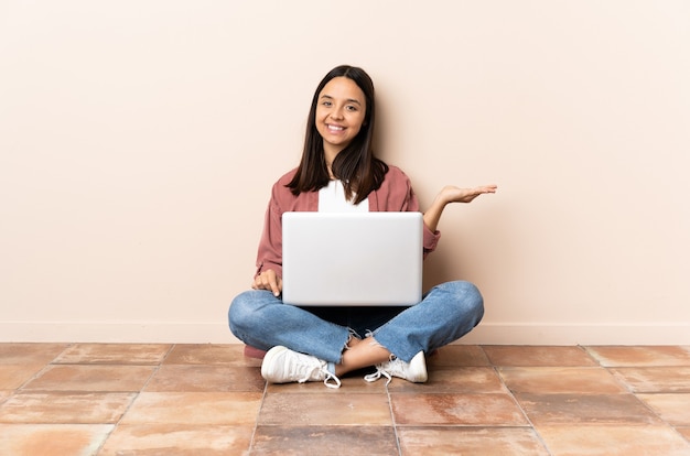 Jeune femme métisse avec un ordinateur portable assis sur le sol tenant copyspace imaginaire sur la paume pour insérer une annonce