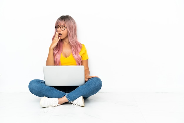 Jeune femme métisse avec un ordinateur portable aux cheveux roses assis sur le sol isolé sur fond blanc bâillant et couvrant la bouche grande ouverte avec la main