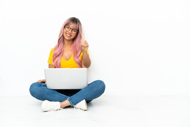 Jeune femme métisse avec un ordinateur portable aux cheveux roses assis sur le sol isolé sur fond blanc montrant et levant un doigt