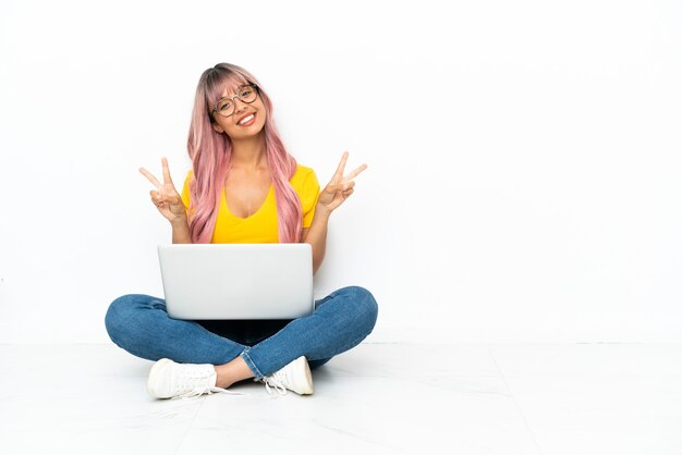 Jeune femme métisse avec un ordinateur portable aux cheveux roses assis sur le sol isolé sur fond blanc montrant le signe de la victoire avec les deux mains