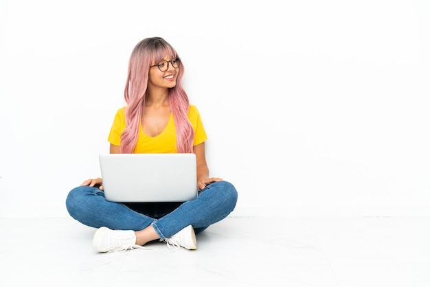 Jeune femme métisse avec un ordinateur portable aux cheveux roses assis sur le sol isolé sur fond blanc regardant sur le côté