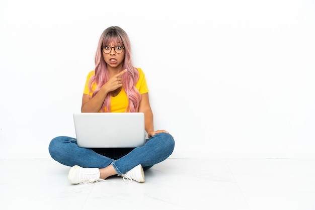 Jeune femme métisse avec un ordinateur portable aux cheveux roses assis sur le sol isolé sur fond blanc surpris et pointant le côté