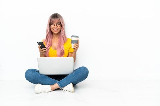 Jeune femme métisse avec un ordinateur portable aux cheveux roses assis sur le sol isolé sur fond blanc tenant du café à emporter et un mobile