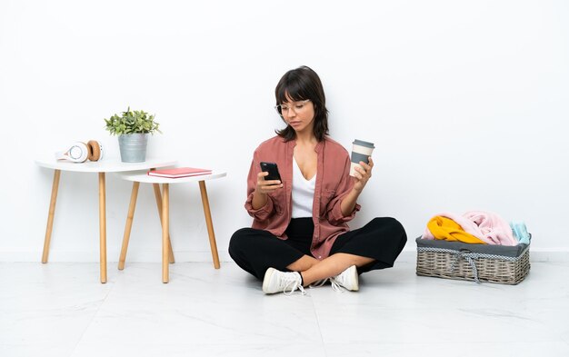 Jeune femme métisse pliant des vêtements assis sur le sol isolé sur fond blanc tenant du café à emporter et un mobile