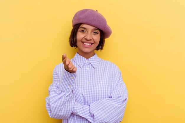 Jeune femme métisse portant un béret isolé sur fond jaune s'étendant la main à la caméra en geste de salutation.