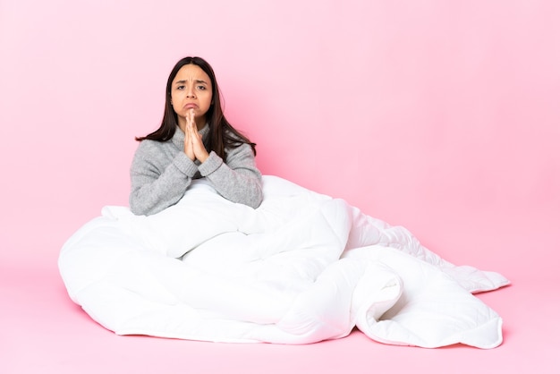 Jeune femme métisse portant pijama assis sur le sol garde la paume ensemble. La personne demande quelque chose