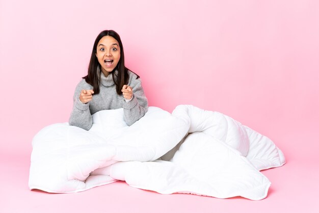 Jeune femme métisse portant pijama assis sur le sol surpris et pointant vers l'avant