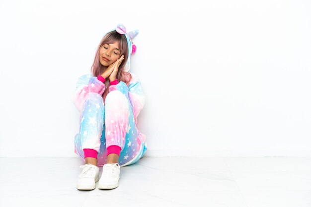 Jeune femme métisse portant un pyjama de licorne assis sur le sol isolé sur fond blanc faisant un geste de sommeil dans une expression adorable