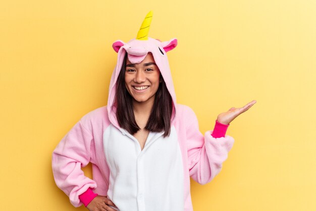 Jeune femme métisse portant un pyjama licorne isolé sur fond jaune montrant un espace de copie sur une paume et tenant une autre main sur la taille.