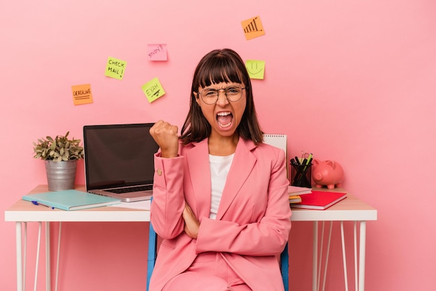 Jeune femme métisse préparant un examen dans la salle tenant un café isolé sur fond rose levant le poing après un concept de vainqueur de la victoire