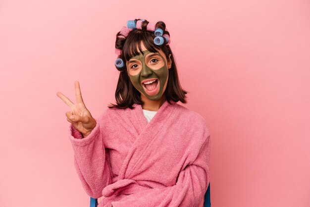 Photo jeune femme métisse se jolie dans une maison isolée sur fond rose joyeuse et insouciante montrant un symbole de paix avec les doigts