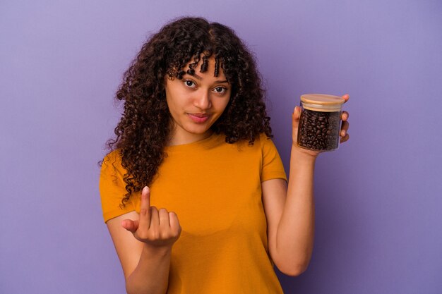 Jeune femme métisse tenant une bouteille de grains de café isolée sur fond violet pointant le doigt vers vous comme si vous vous invitiez à vous rapprocher.