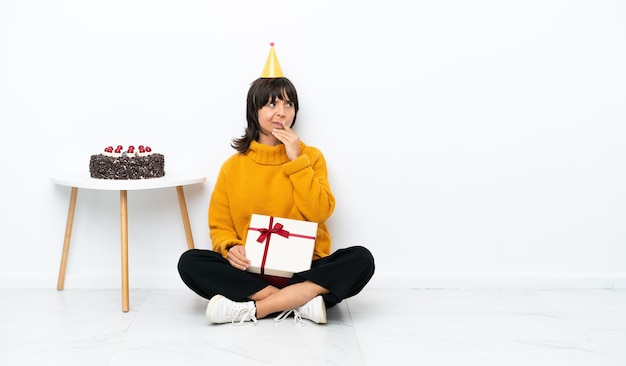 Jeune femme métisse tenant un cadeau assis sur le sol isolé sur fond blanc ayant des doutes et avec une expression de visage confuse