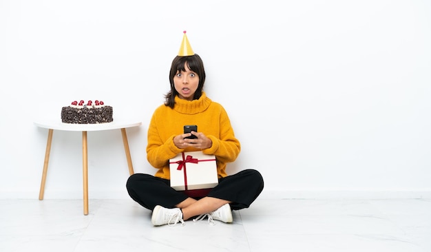 Jeune femme métisse tenant un cadeau assis sur le sol isolé sur fond blanc surpris et envoyant un message