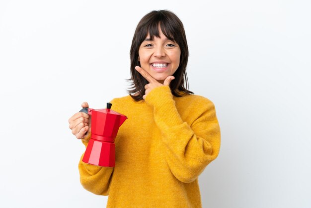 Jeune femme métisse tenant une cafetière isolé sur fond blanc heureux et souriant