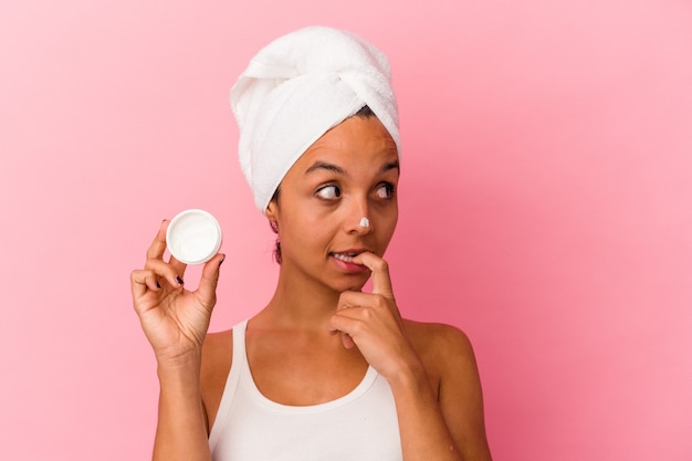 Jeune femme métisse tenant une crème pour le visage isolée sur fond rose détendue en pensant à quelque chose en regardant un espace de copie.