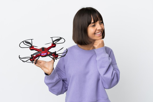 Jeune femme métisse tenant un drone isolé sur fond blanc regardant sur le côté et souriant