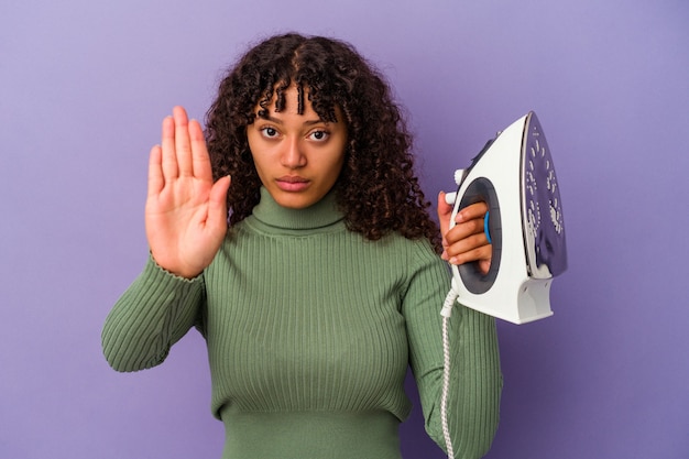 Jeune femme métisse tenant un fer isolé sur fond violet debout avec la main tendue montrant un panneau d'arrêt, vous empêchant.