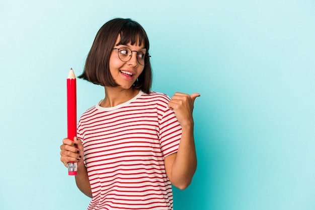 Jeune femme métisse tenant un gros crayon isolé sur des points de fond bleu avec le pouce loin, riant et insouciant.