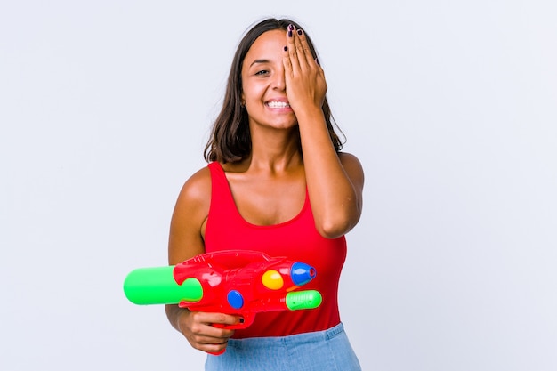 Jeune femme métisse tenant un pistolet à eau isolé s'amusant couvrant la moitié du visage avec la paume.