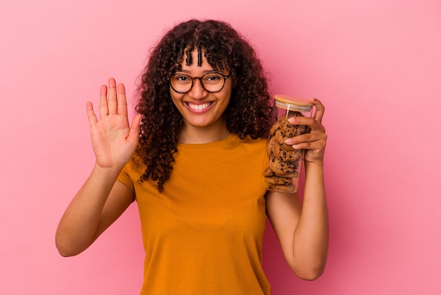 Jeune femme métisse tenant un pot de cookies isolé sur fond rose souriant joyeux montrant le numéro cinq avec les doigts.