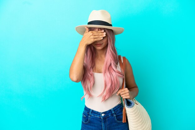 Jeune femme métisse tenant un sac de plage avec pamela isolé sur fond bleu couvrant les yeux à la main. Je ne veux pas voir quelque chose