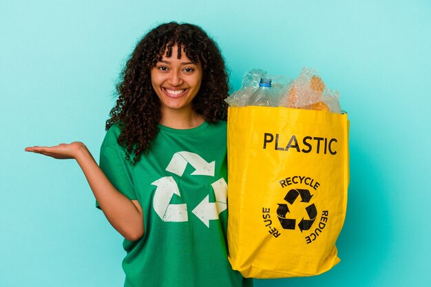 Jeune femme métisse tenant un sac en plastique recyclé isolé sur fond bleu montrant un espace de copie sur une paume et tenant une autre main sur la taille.