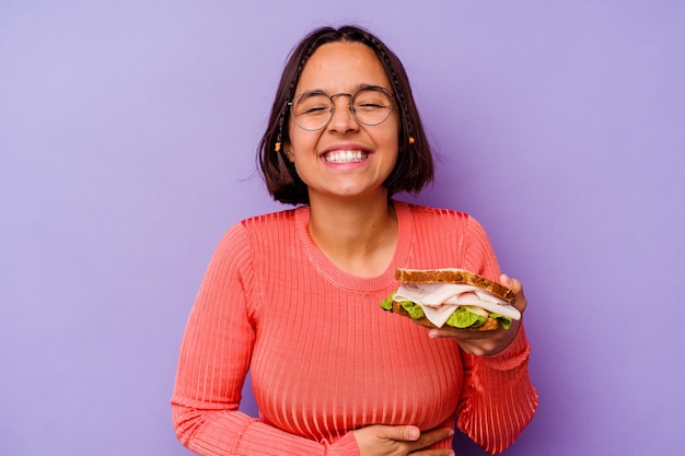 Jeune femme métisse tenant un sandwich isolé sur fond violet en riant et en s'amusant.