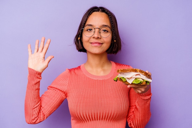 Jeune femme métisse tenant un sandwich isolé sur fond violet souriant joyeux montrant le numéro cinq avec les doigts.