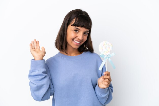 Jeune femme métisse tenant une sucette isolée sur fond blanc saluant avec la main avec une expression heureuse