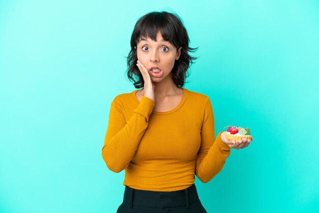 Jeune femme métisse tenant une tartelette isolée sur fond bleu avec une expression faciale surprise et choquée