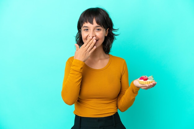 Jeune femme métisse tenant une tartelette isolée sur fond bleu heureuse et souriante couvrant la bouche avec la main