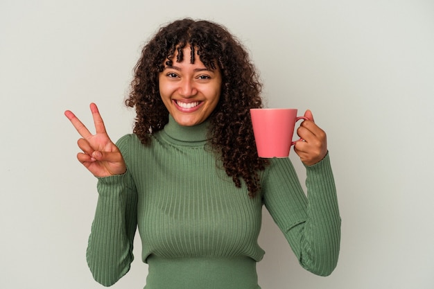 Jeune femme métisse tenant une tasse isolée sur fond blanc montrant le numéro deux avec les doigts.