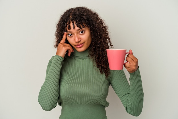 Jeune femme métisse tenant une tasse isolée sur fond blanc pointant le temple avec le doigt, pensant, concentrée sur une tâche.