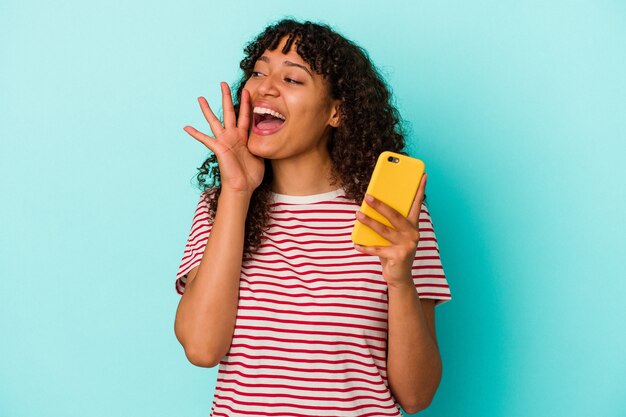 Jeune femme métisse tenant un téléphone portable isolé sur fond bleu criant et tenant la paume près de la bouche ouverte.