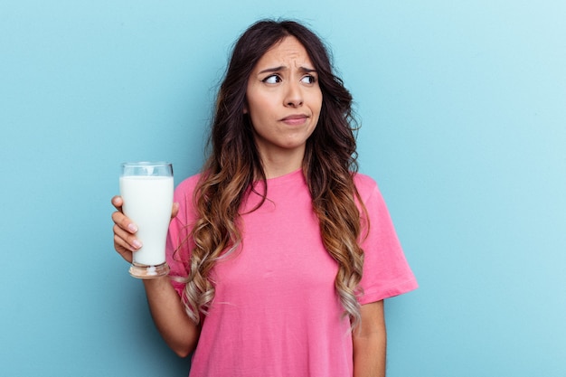 Jeune femme métisse tenant un verre de lait isolé sur fond bleu confus, se sent dubitative et incertaine.